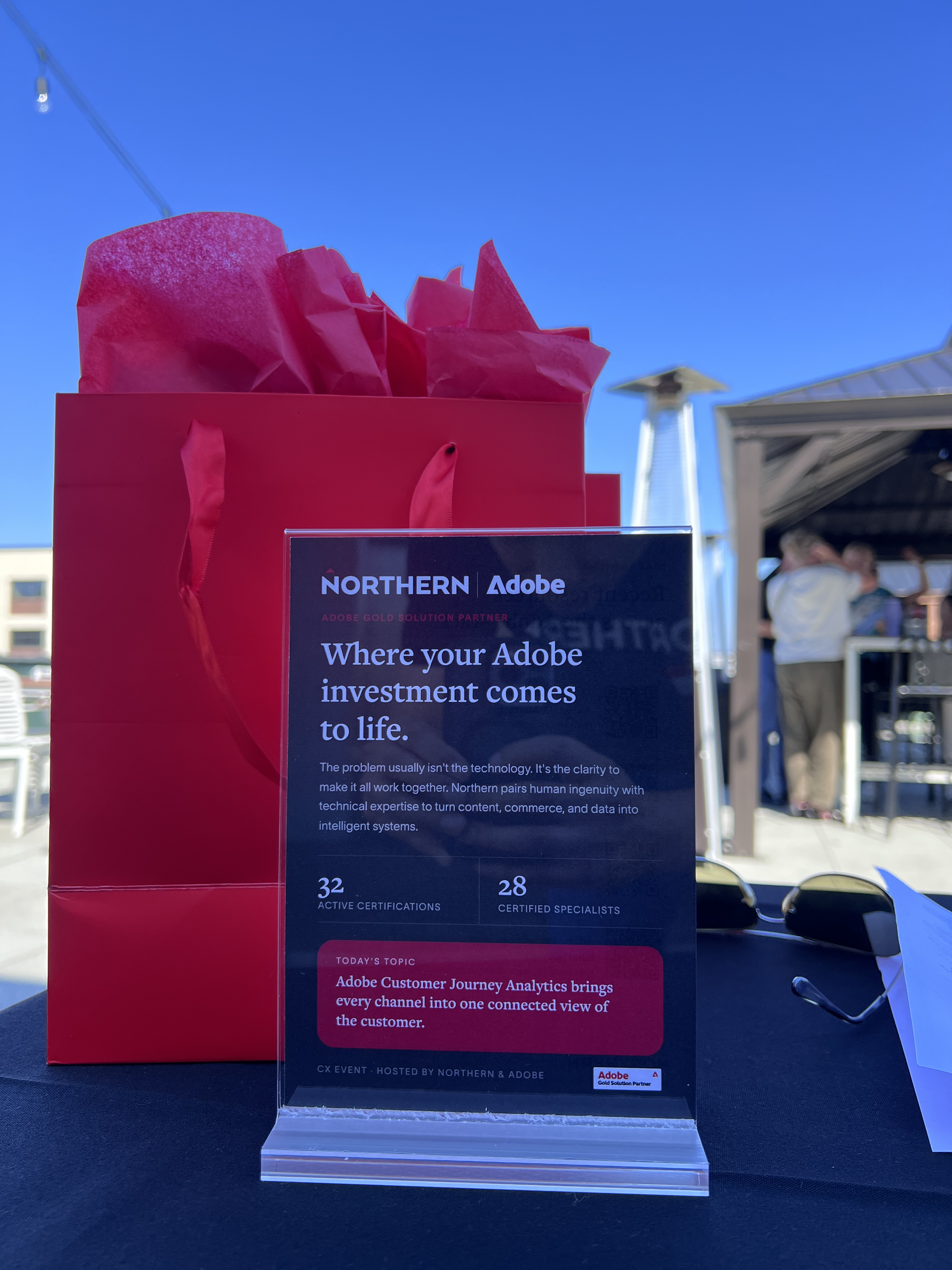 Northern and Adobe table card reading "Where your Adobe investment comes to life" beside a red gift bag on a sunny rooftop