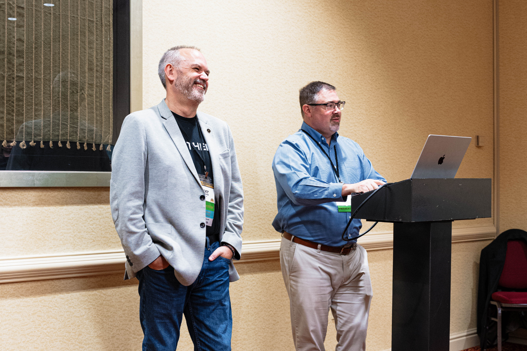 Jay Ménard from Northern stands smiling beside Hal Legg, Director of PR at SUNY Niagara, who is presenting from a podium with a laptop, at the NCMPR conference.