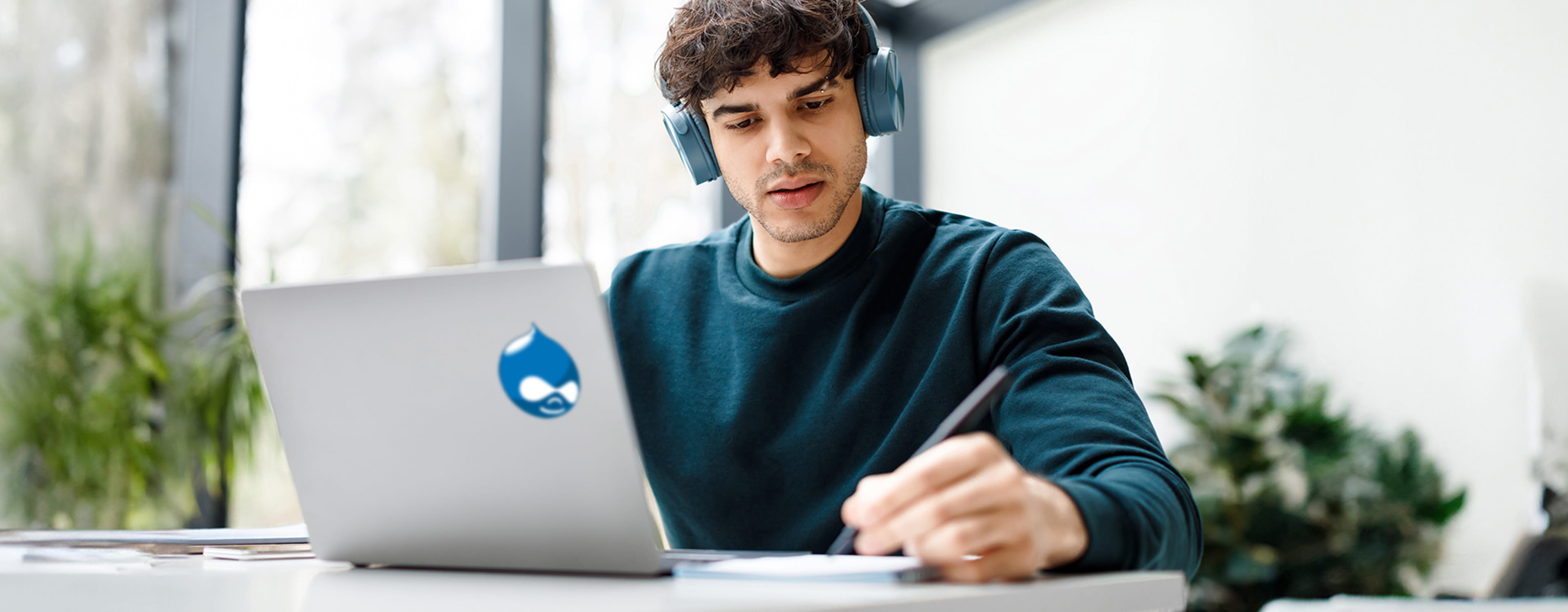Man with headphones on looking at laptop with Drupal logo on the back of it