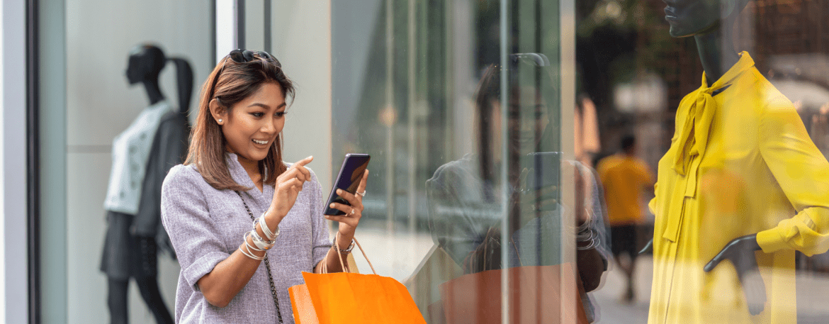 Smiling shopper checking her phone while window shopping at a storefront, holding a shopping bag
