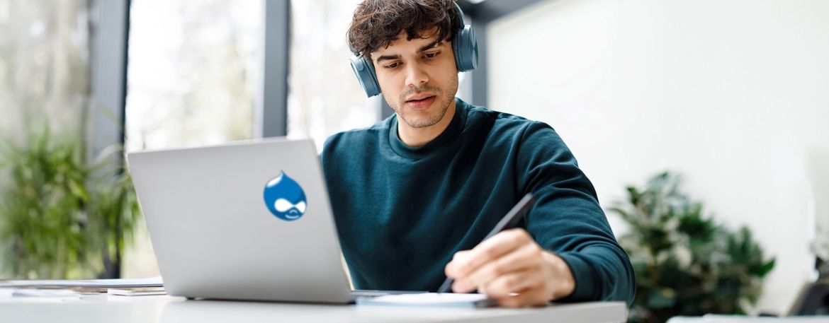 Man with headphones on looking at laptop with Drupal logo on the back of it