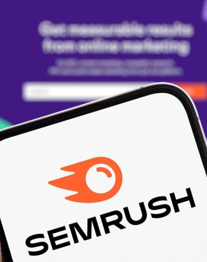 The Semrush logo on a phone screen, held in front of the Semrush website in the background.