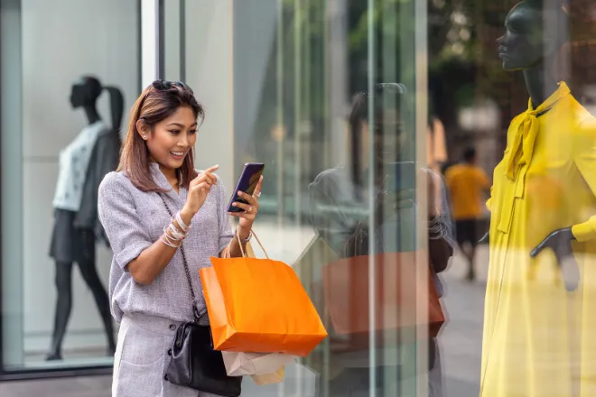 Smiling shopper checking her phone while window shopping at a storefront, holding a shopping bag