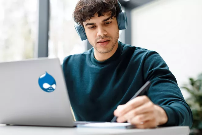 Man with headphones on looking at laptop with Drupal logo on the back of it