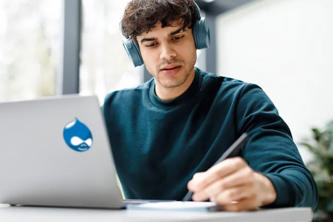 Man with headphones on looking at laptop with Drupal logo on the back of it
