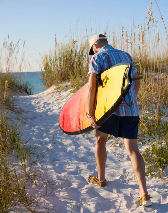 Man carrying a surfboard along a sandy beach path wearing Quarks sandals