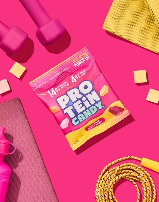 Pink pouch of protein candy on a pink background with weights, a yoga mat, and a water bottle